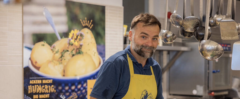 Ein glücklicher Mitarbeiter steht hinter der Theke im Bio-Markt Vorwerk Podemus und lächelt.